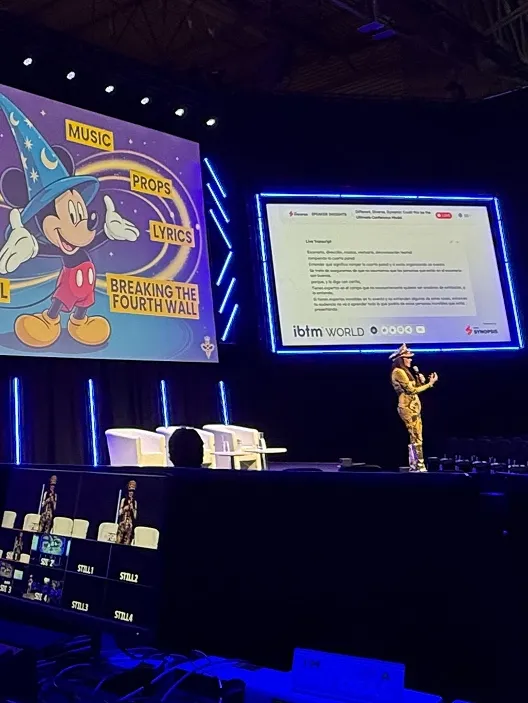 Speaker dressed in patterned outfit with a hat presenting on stage with two large screens, one showing a cartoon Mickey Mouse with labels 'MUSIC,' 'PROPS,' 'LYRICS,' and 'BREAKING THE FOURTH WALL,' and the other displaying a text slide labeled 'ibtm WORLD.'