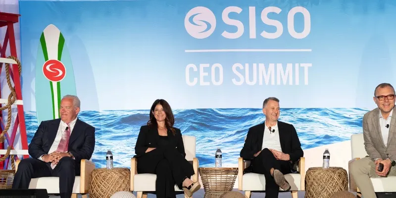 Four panelists seated on white chairs on stage at the SISO CEO Summit with ocean-themed backdrop and surfboard.