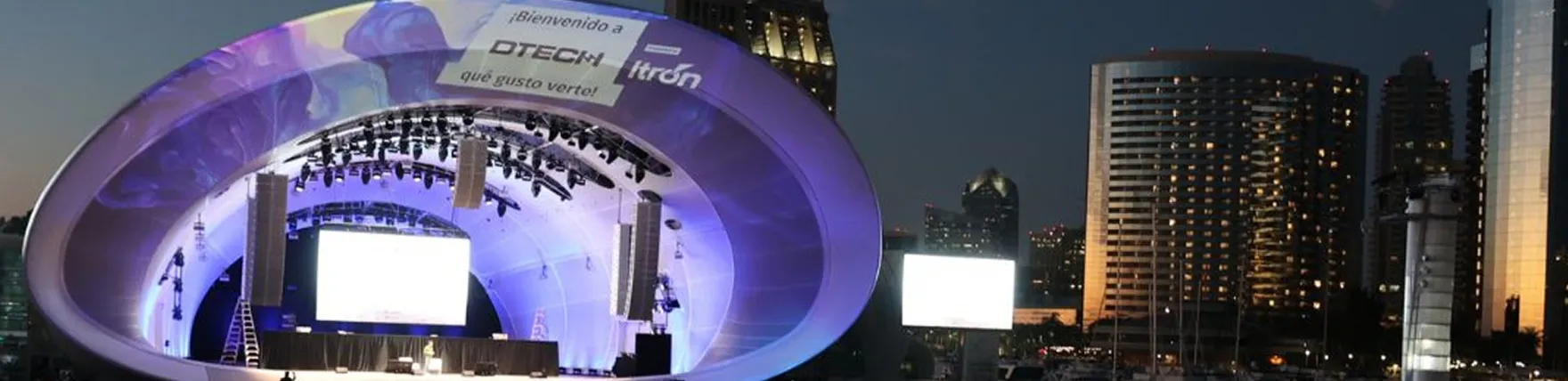 Outdoor stage with curved purple-lit canopy at dusk with city buildings in the background.