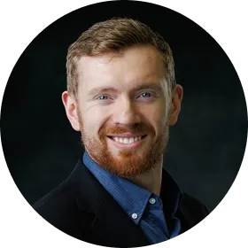 Smiling man with red hair and beard wearing a dark blazer and blue shirt against a dark background.