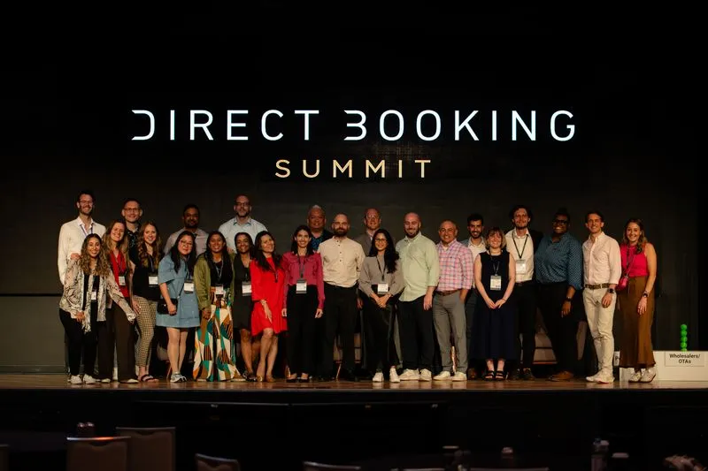Group of 20 diverse people standing on stage in front of a 'Direct Booking Summit' sign, posing for a photo.
