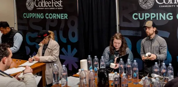 Four people at a CoffeeFest cupping corner booth, tasting and evaluating coffee samples with water bottles and cups on the table.
