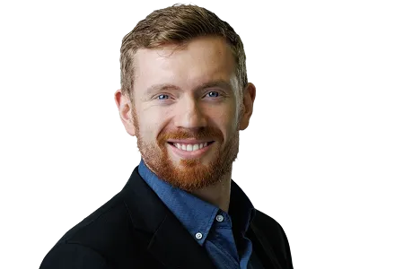 Smiling man with red hair and beard wearing a dark blazer and blue shirt on a transparent background.