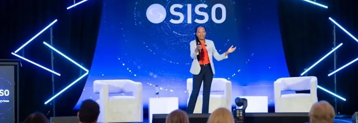 Speaker presenting on stage with SISO logo on blue background and empty white chairs on each side.