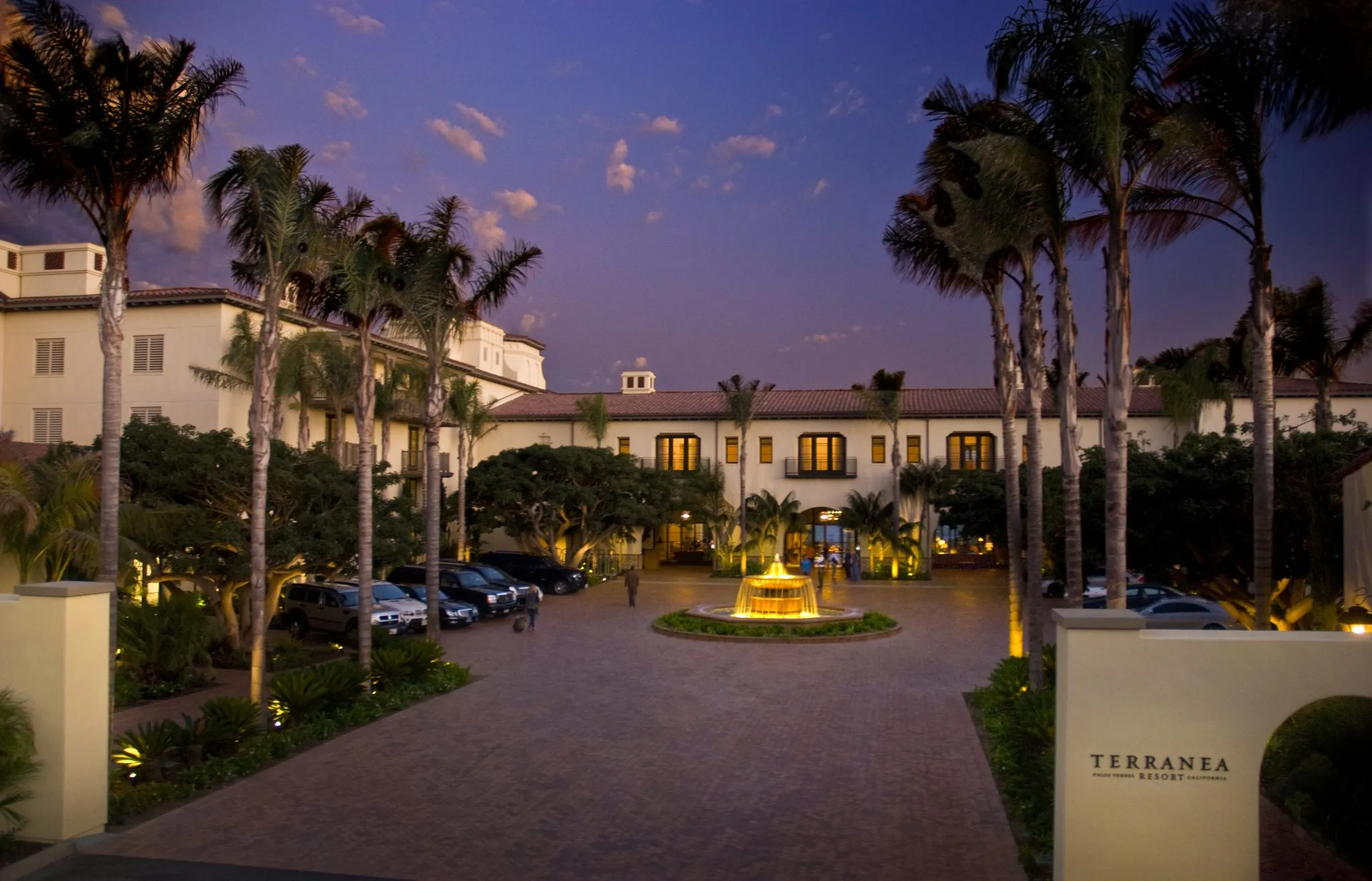 Palm-lined entrance of Terranea Resort at dusk with a lit fountain and parked cars.