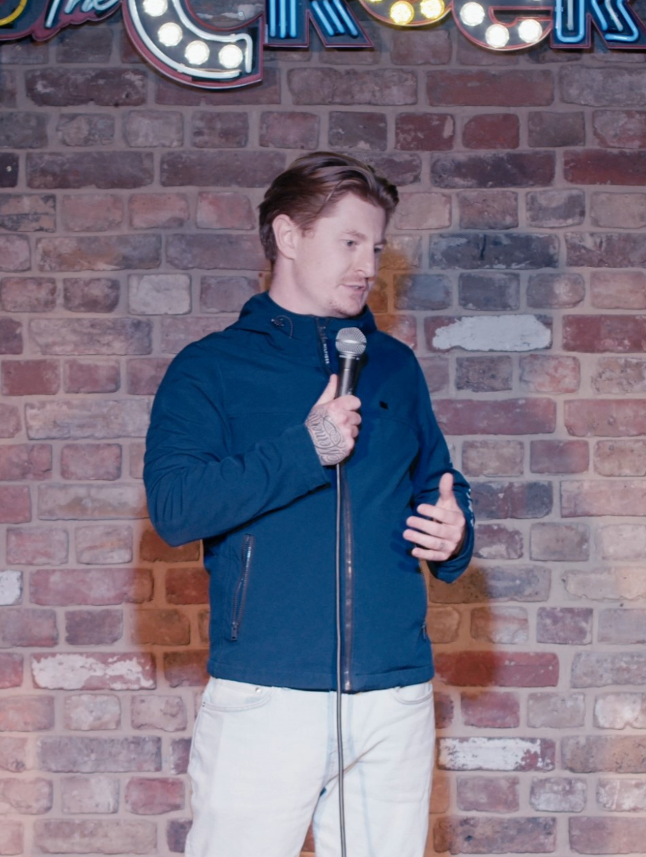 Comedian Tom Nestor in a blue jacket holds a microphone, speaking animatedly. They're on a stage with a brick wall backdrop and partly visible illuminated letters. This photo is used to promote an upcoming performance at Comedy Freaks Kings Cross.