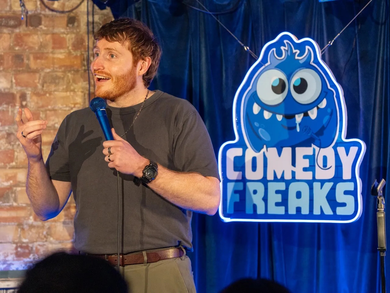 British Comedian Ali Woods on stage with a microphone, gesturing expressively. Background features a brick wall and a neon sign with "Comedy Freaks" and a cartoon monster at a London comedy club.