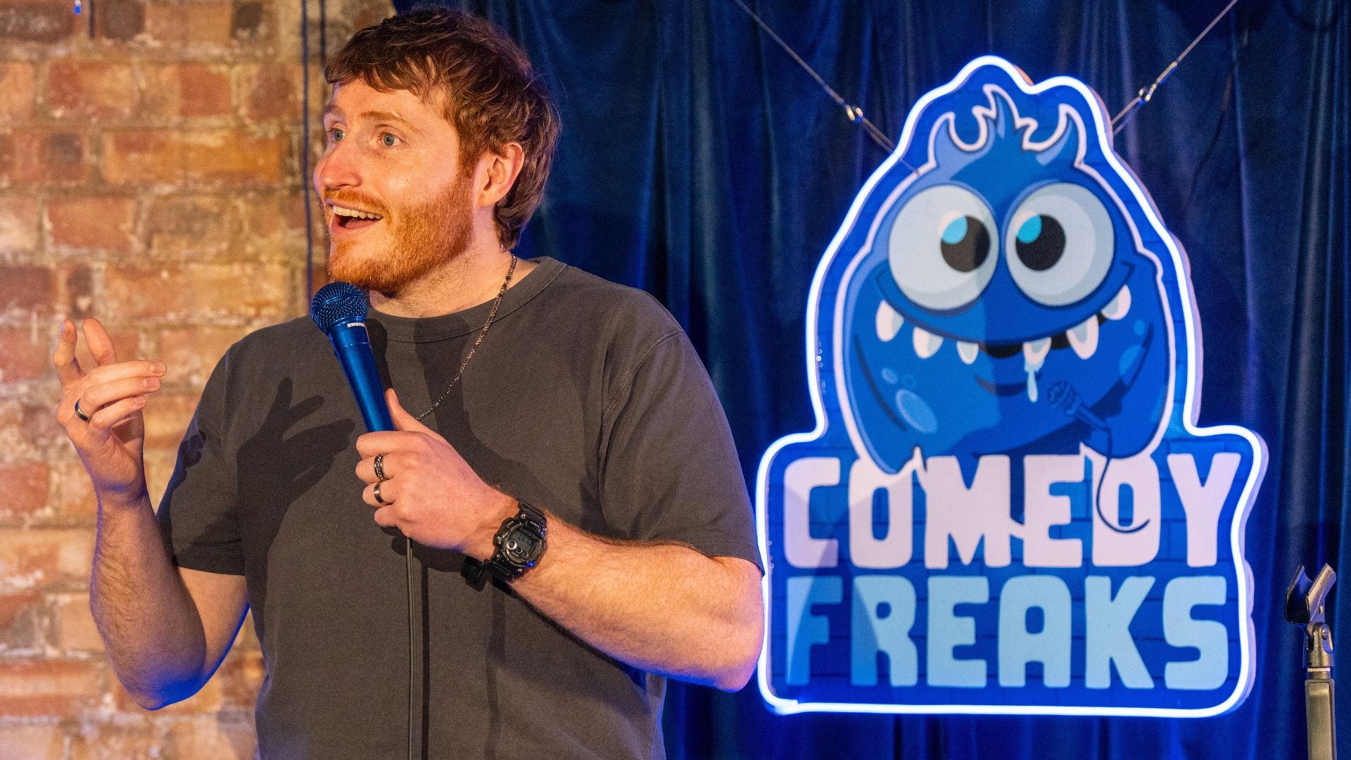 Comedian Ali Woods performing at comedy club London, holding microphone close to face, wearing dark t-shirt against exposed brick wall with blue stage lighting and Comedy Freaks logo backdrop