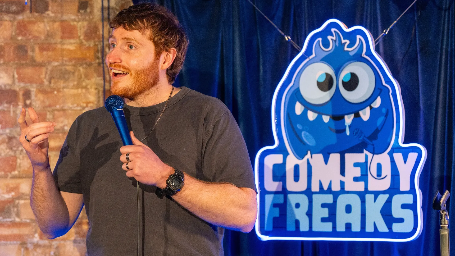 Comedian Ali Woods performing at comedy club London, holding microphone close to face, wearing dark t-shirt against exposed brick wall with blue stage lighting and Comedy Freaks logo backdrop