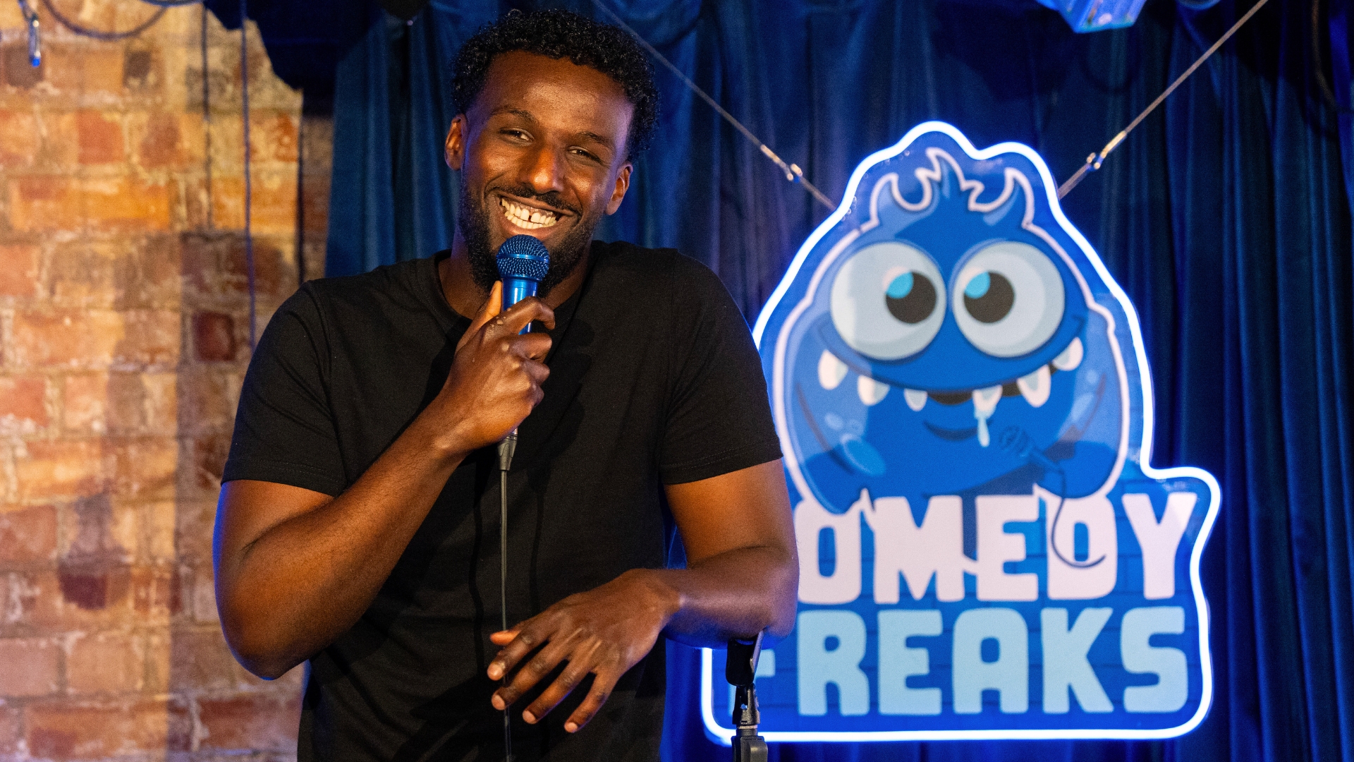 Bamph, a male comedian with a bright smile, holds a microphone on stage in front of a brick wall and the glowing Comedy Freaks logo, exuding energetic and unpredictable vibes at London premier destination for cutting-edge stand up comedy.