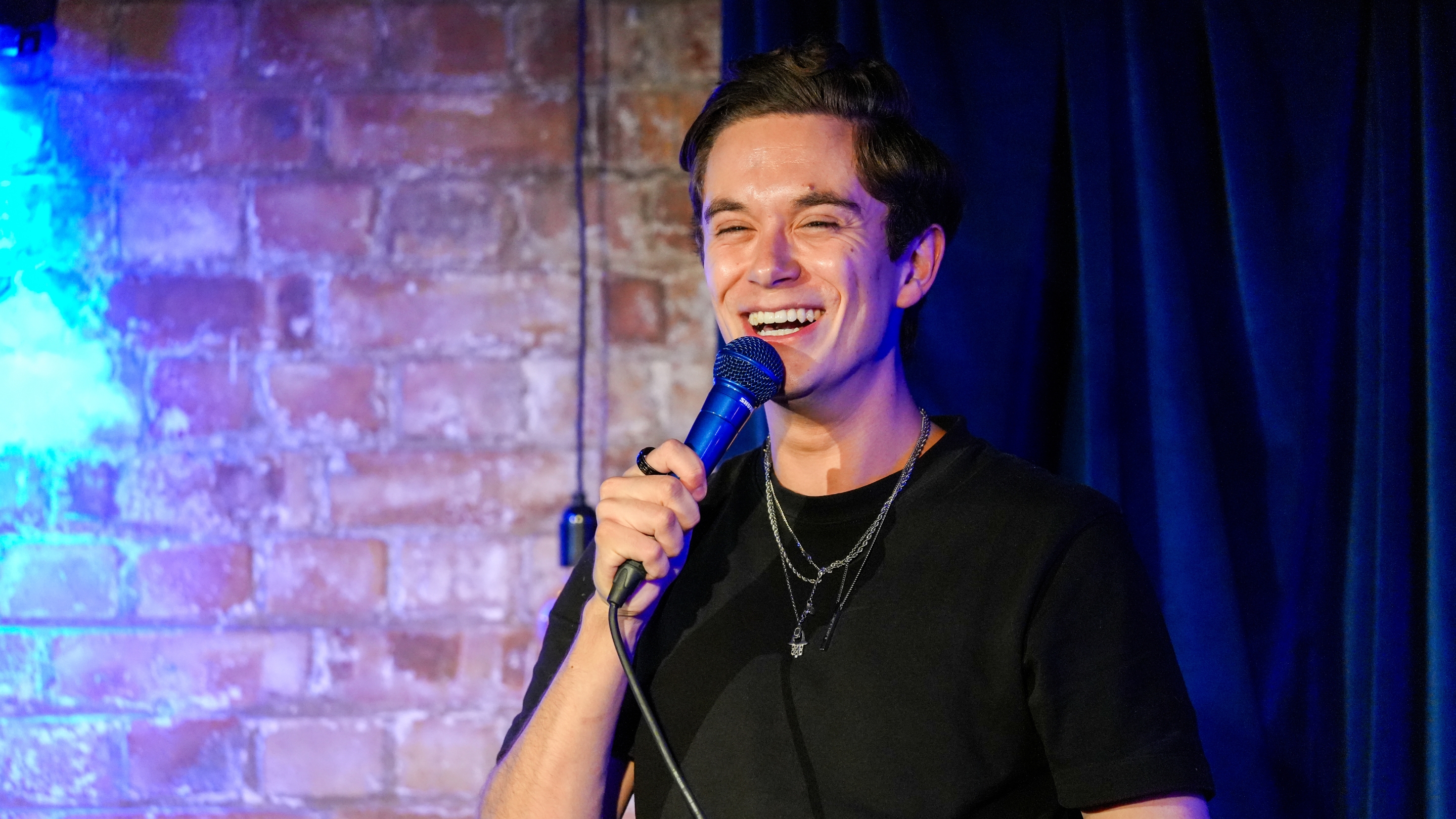 Jomi Cruz, a male comedian with dark hair, laughs while holding a microphone on stage against a brick wall with blue lighting, delivering an energetic performance at Comedy Freaks - The Most Unique Comedy Club in London.