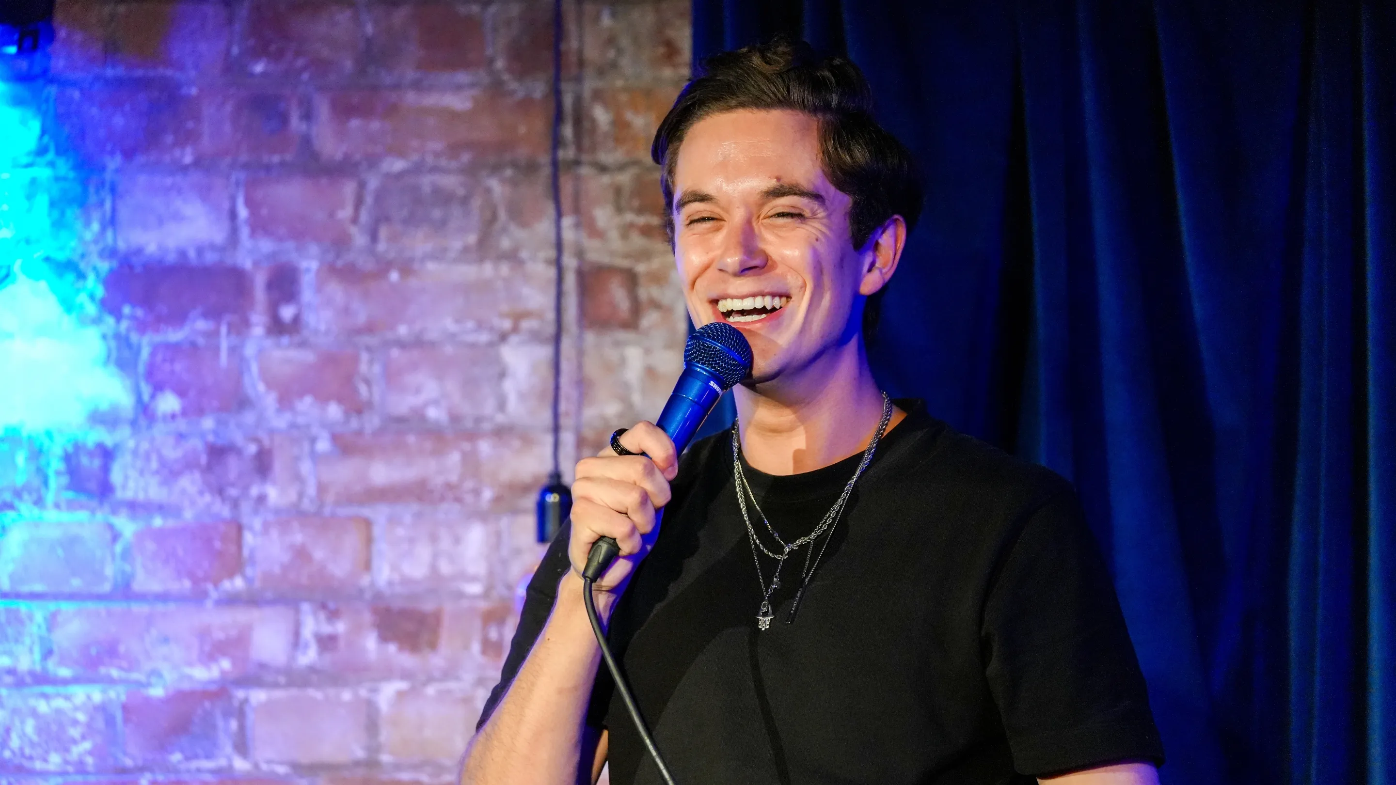Jomi Cruz, a male comedian with dark hair, laughs while holding a microphone on stage against a brick wall with blue lighting, delivering an energetic performance at Comedy Freaks - The Most Unique Comedy Club in London.