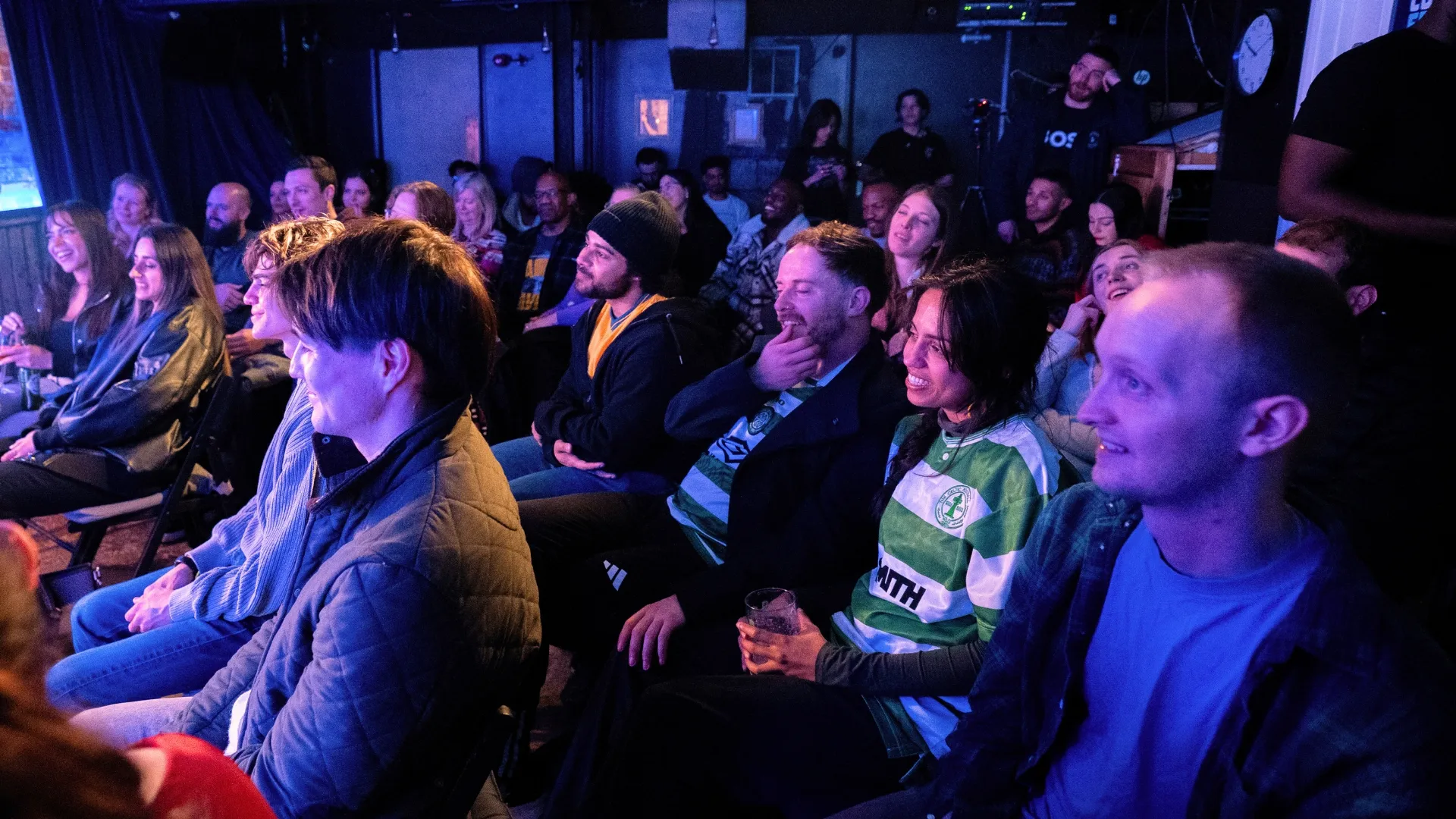 Engaged audience, smiling and attentive, seated in a dimly lit club with brick walls and blue lighting, enjoying a captivating live stand-up show at Comedy Freaks, a unique comedy show London.