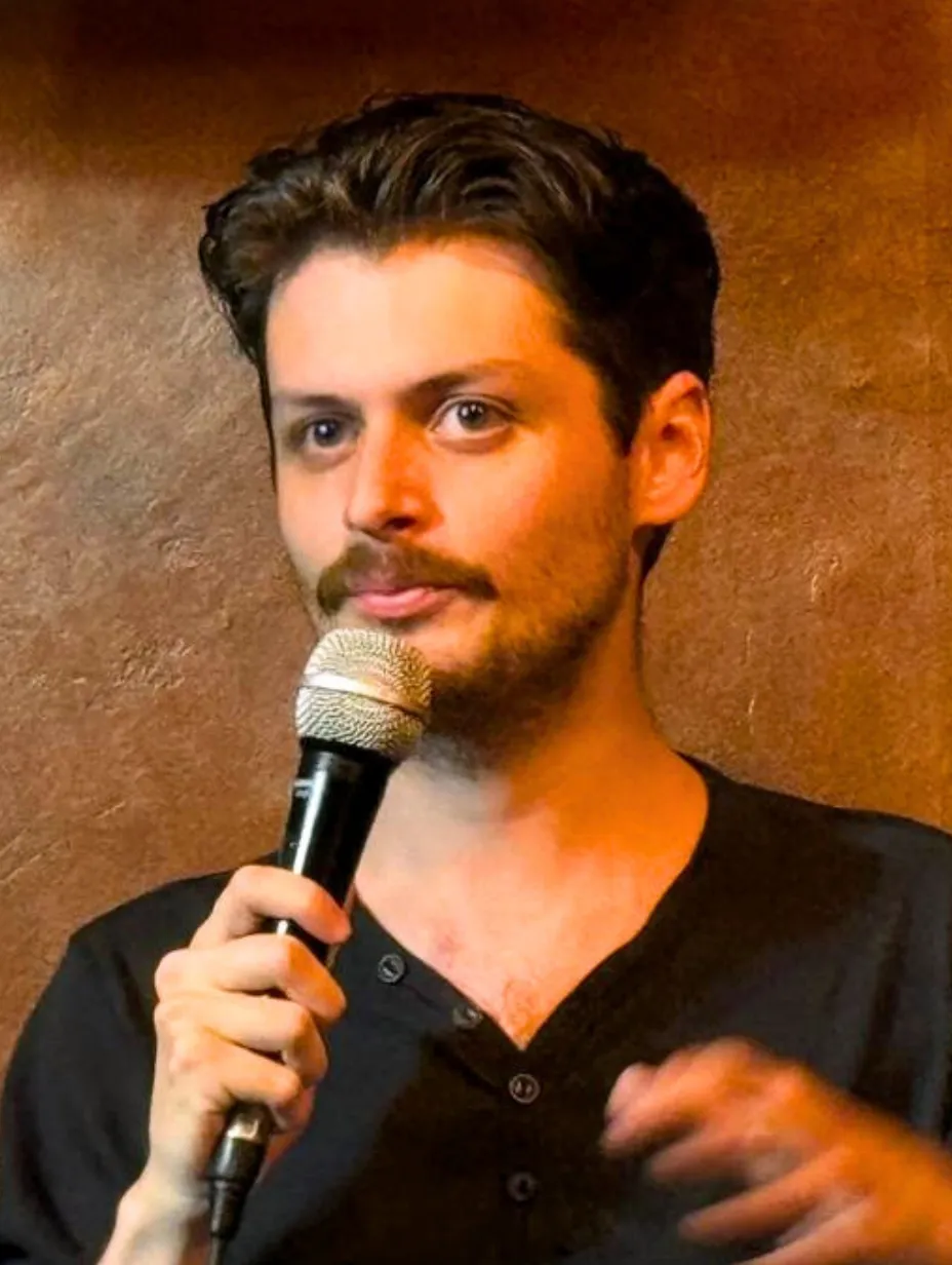 Will Burke, a high-energy male comedian from the Channel Islands, with dark hair and a moustache, holds a microphone and gestures on stage against a warm, textured wall. He's delivering stand-up at a Comedy Freaks comedy show London.