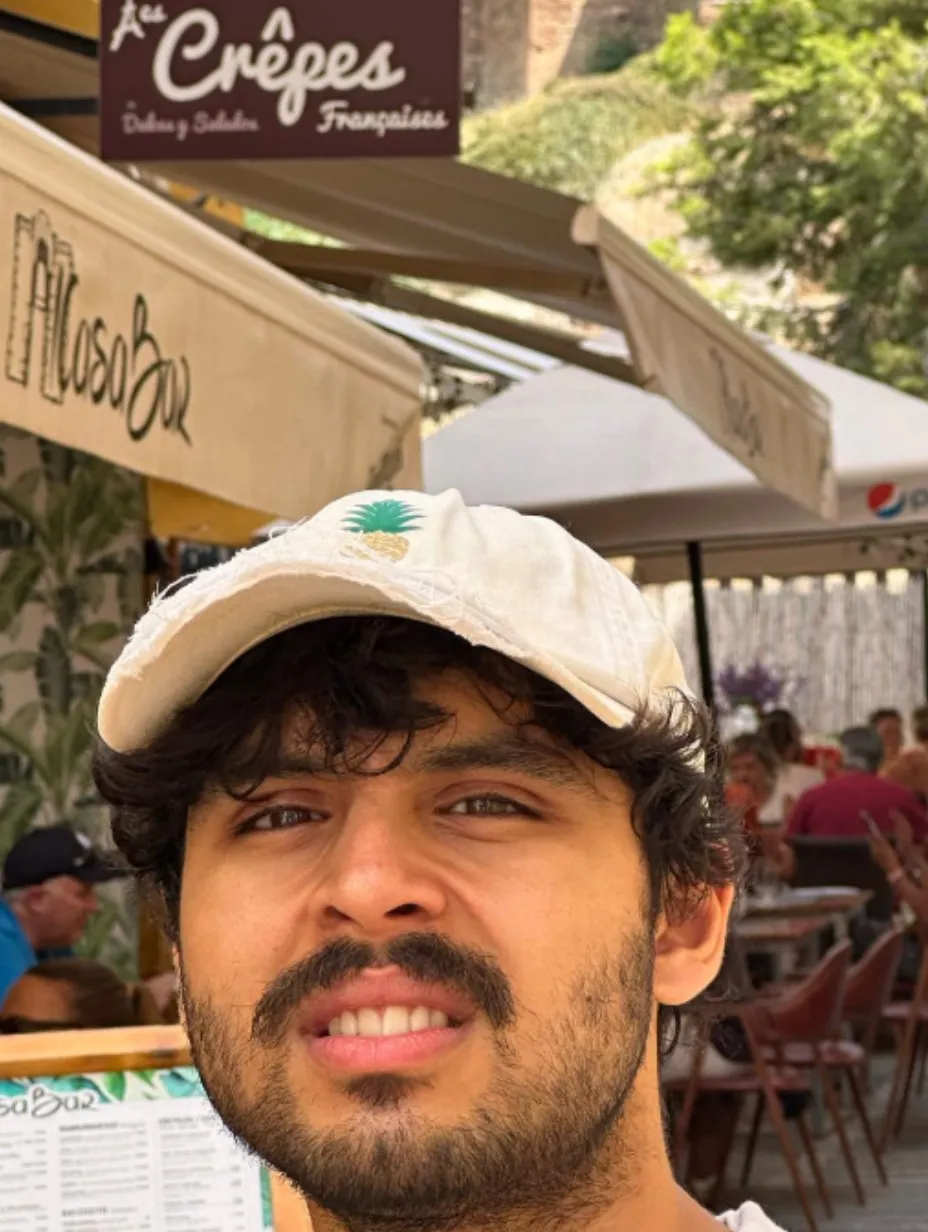 Abhi Wagle, a male comedian with a beard and a white cap, smiles engagingly at the camera, with a restaurant or cafe setting and outdoor seating in the background. He's ready to deliver his sharp observational comedy at a Comedy Freaks comedy show London