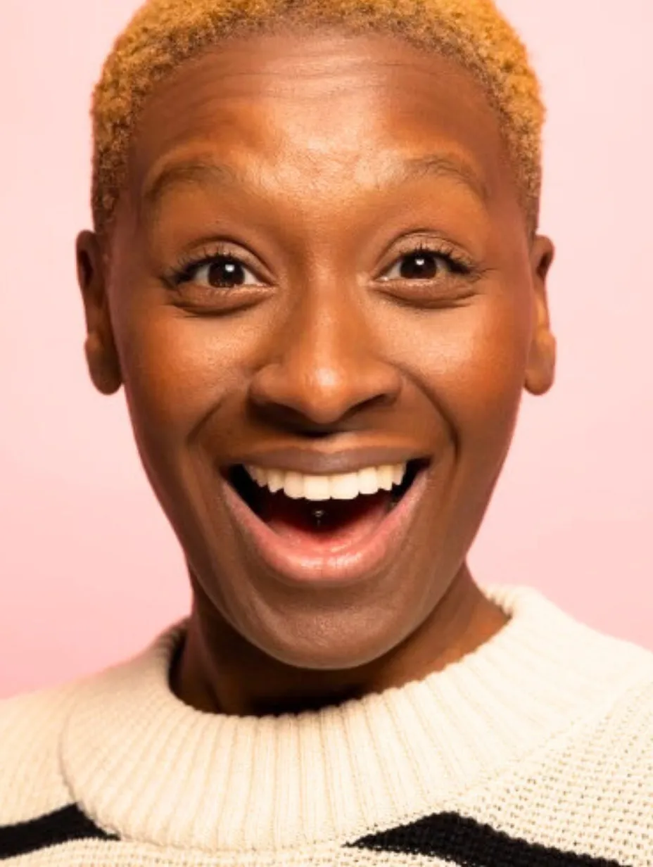 Chantel Nash, a female comedian with short blonde hair, wide eyes, and an open-mouthed smile, wears a cream and black striped jumper against a pink background, exuding high energy for a Comedy Freaks comedy show London