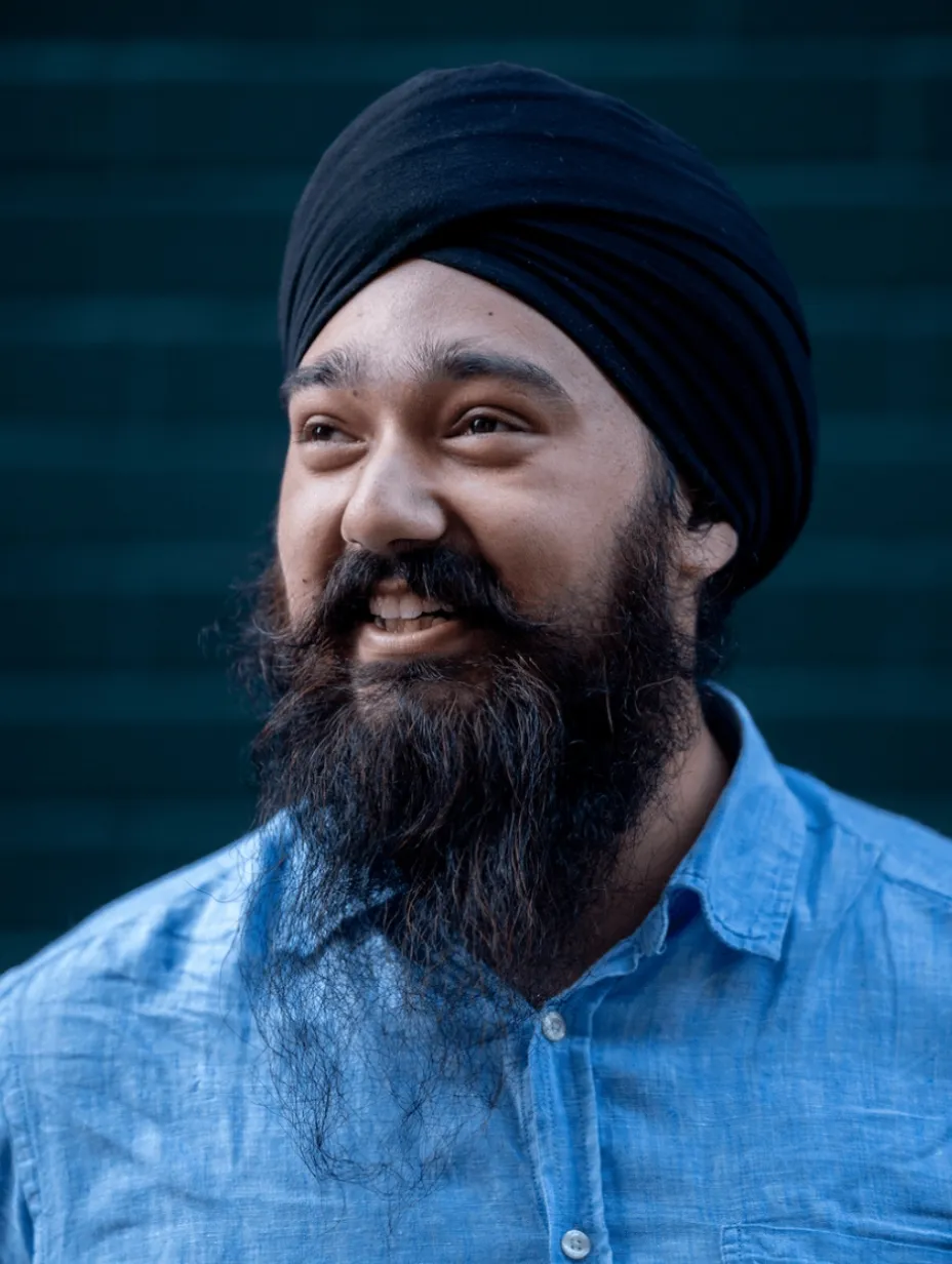 Smiling Daman Bamrah with a full beard and black turban, wearing a light blue shirt. The comedian is set against a dark, blurred background, conveying warmth and positivity.