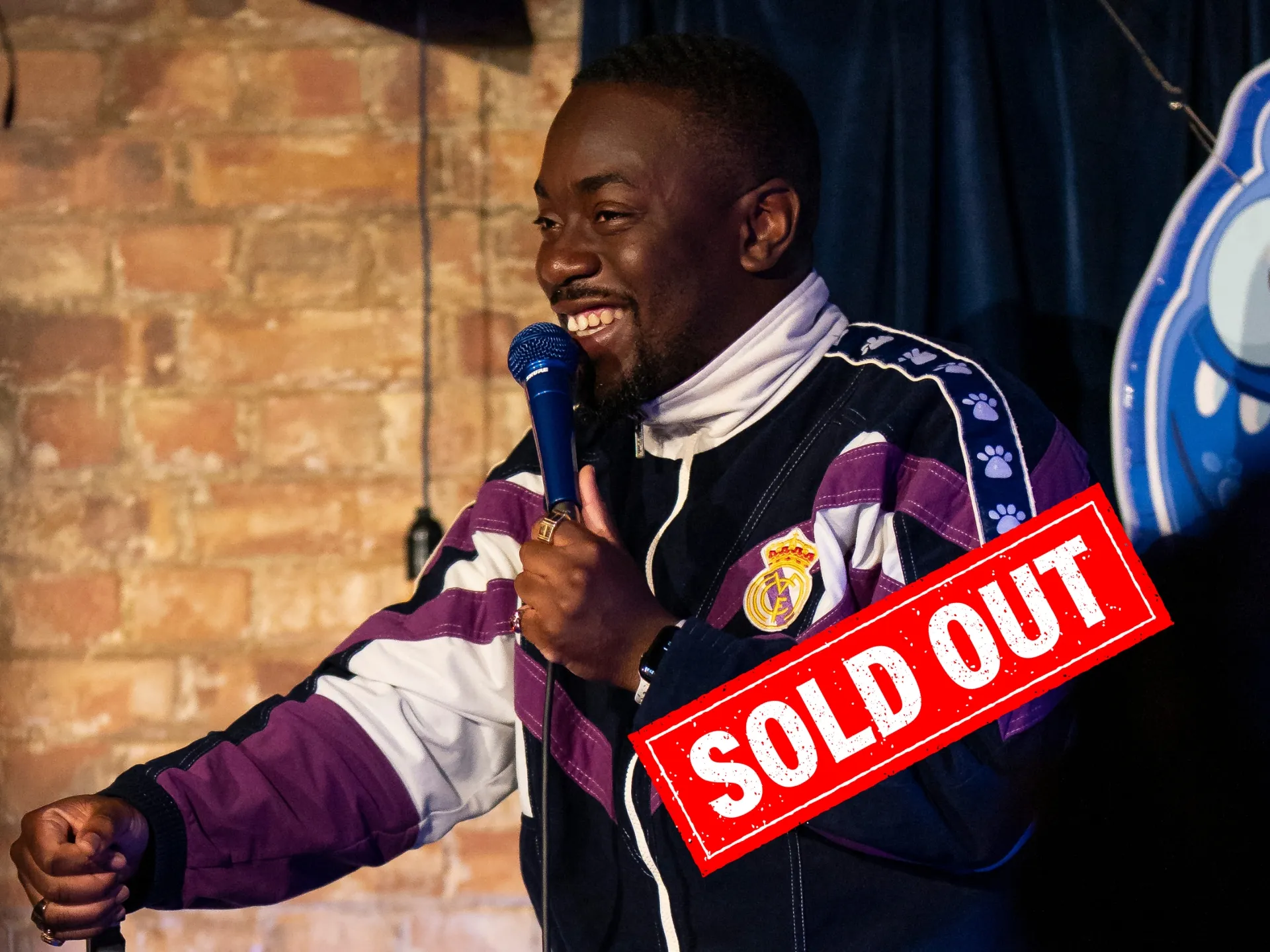 Andrew Mensah, a male black comedian, smiles while holding a microphone and gesturing with his other hand, on stage with a brick wall backdrop and the glowing blue Comedy Freaks logo, delivering energetic stand-up comedy at our London comedy venue.