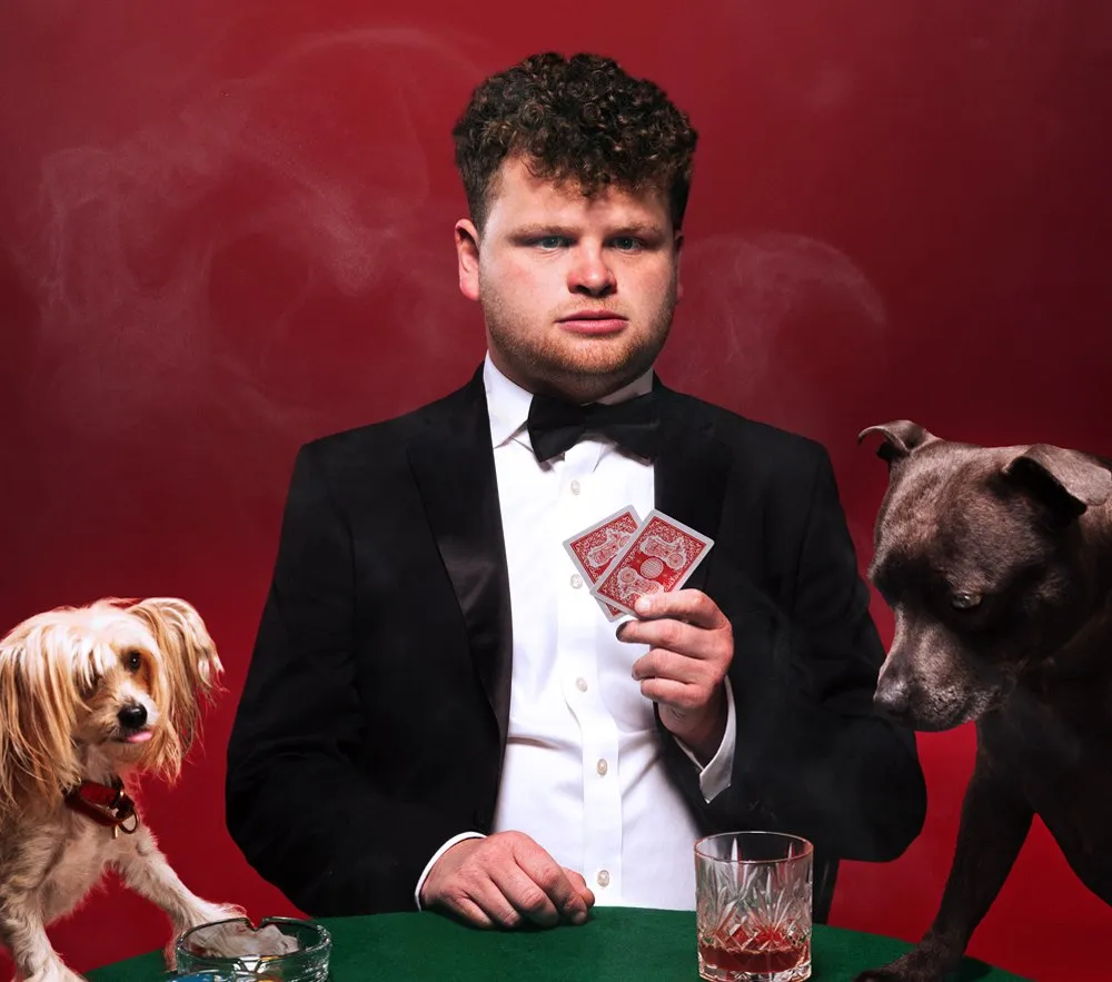 BBC New Comedian of the Year winner Dan Tiernan in a tuxedo, holding a pair of playing cards with two dogs for his Edinburgh Fringe show, "All In."