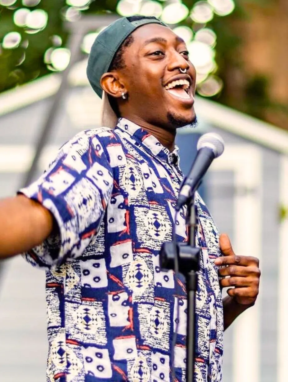 Darnell Jesse, a male comedian, smiles widely and holds a microphone, wearing a backwards baseball cap and a patterned short-sleeve shirt.