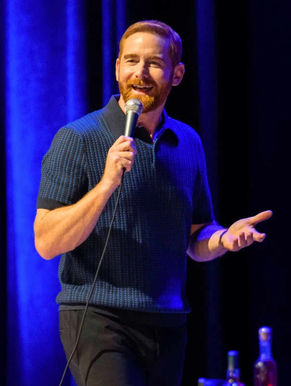 American comedian Andrew Santino performing stand-up comedy on a stage in London, holding a microphone and gesturing to the audience.