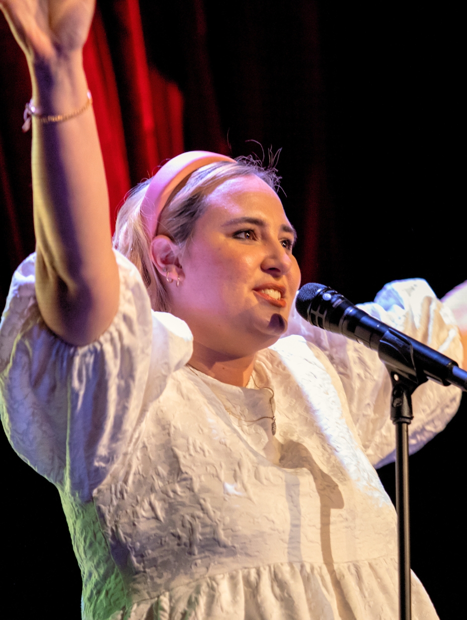 Maddie HW, a female comedian, smiles brightly on stage with one hand raised, wearing a white top and a headband, ready to deliver her energetic and quirky humour at a Comedy Freaks comedy show London.