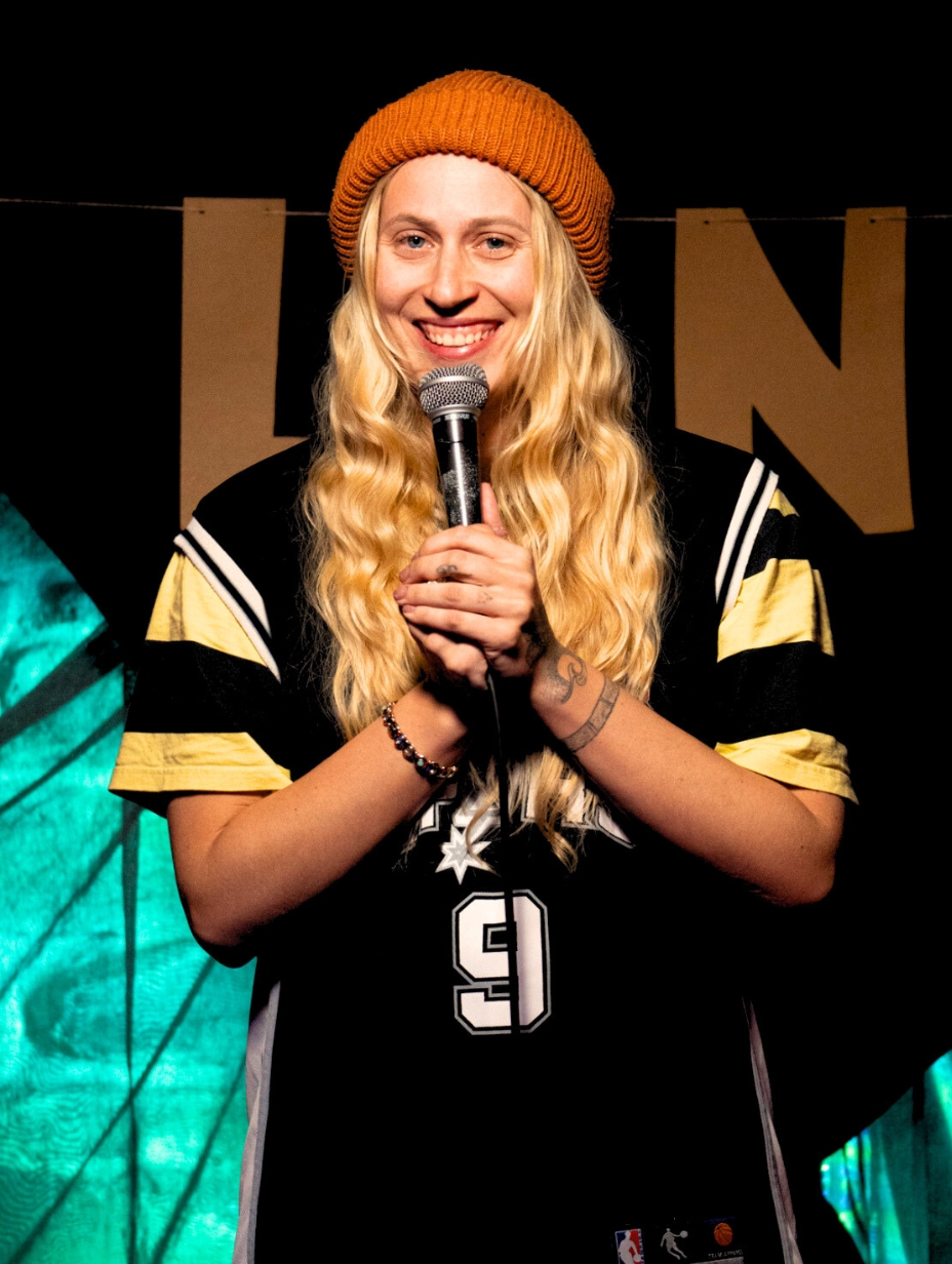Liisel Nelis, a female comedian, smiles broadly while holding a microphone, wearing a bright orange beanie and a black and yellow basketball jersey. Her passionate stage presence is a perfect fit for a Comedy Freaks comedy show London.