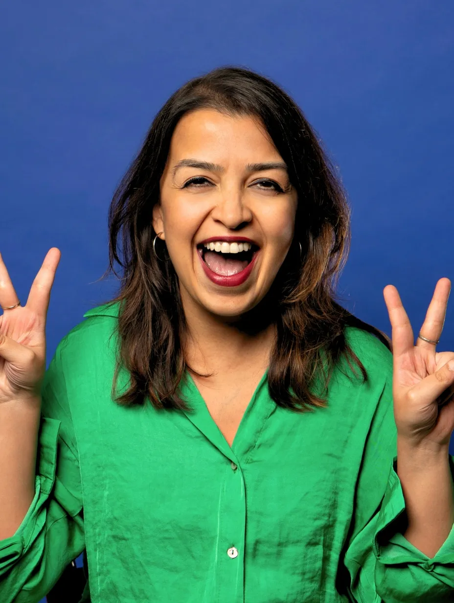 Monica Chatterjee, a female comedian, smiles widely with her mouth open and holds up two peace signs, wearing a bold green shirt against a blue background, representing her bold, cheeky, and chaotic energy at a Comedy Freaks comedy show London.
