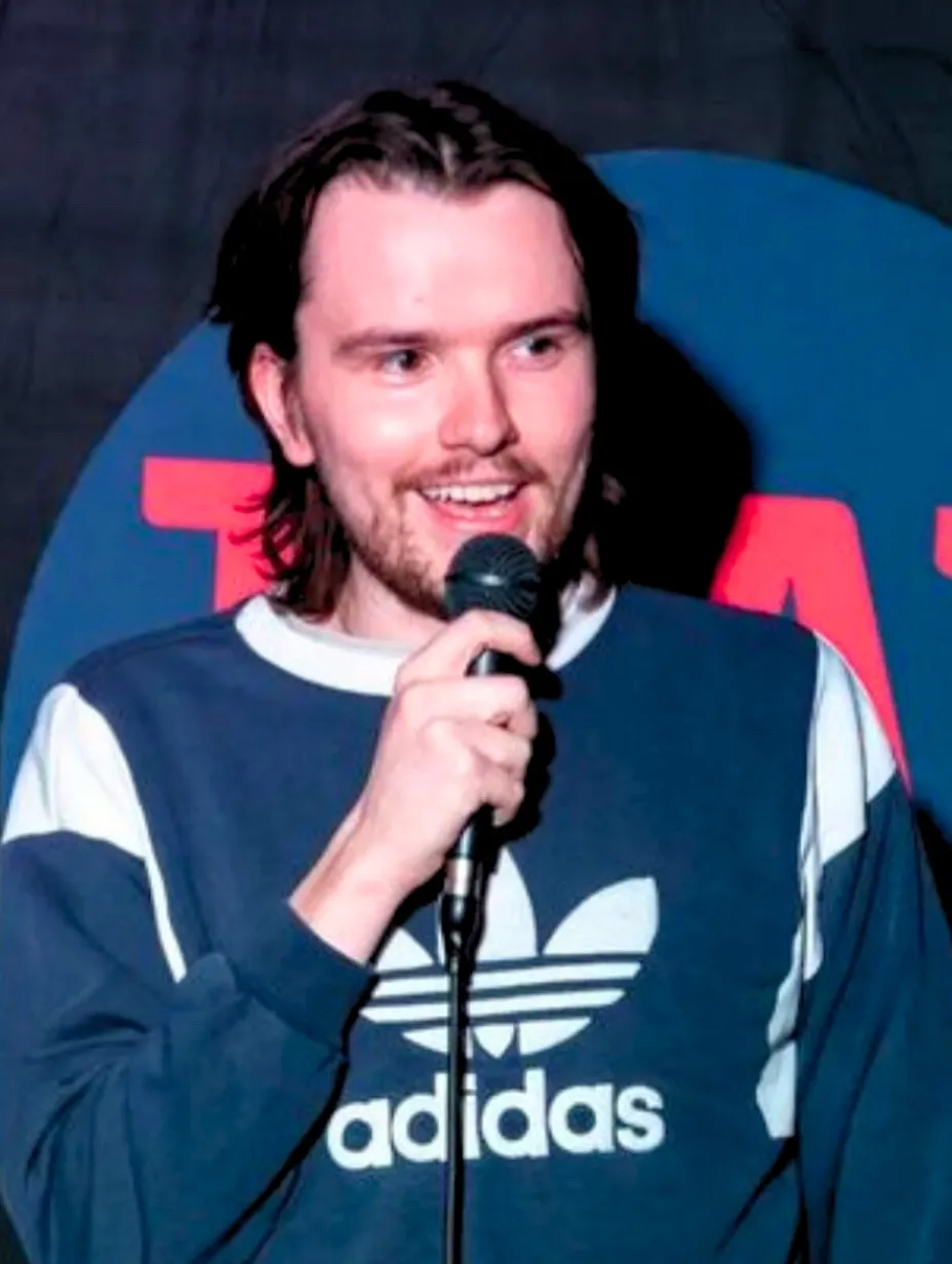 James Pendleton, a male comedian with longish hair and a beard, holds a microphone and smiles while wearing a dark blue Adidas sweatshirt, ready to deliver his sharp observational humour and deadpan delivery at a Comedy Freaks comedy show London.