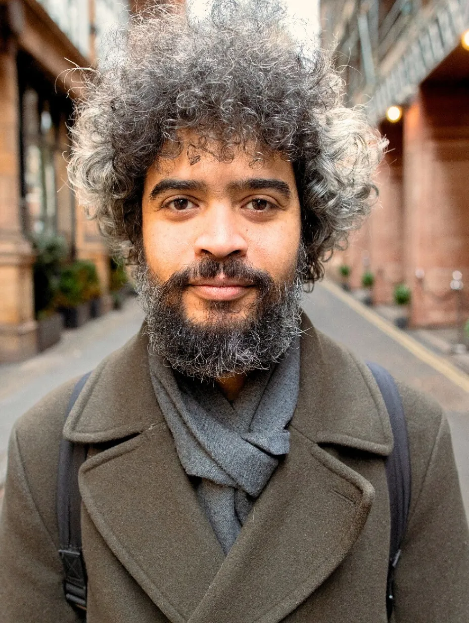 Masud Milas, a male comedian with a beard and curly hair, looks directly at the camera while wearing a dark coat and scarf. He's a prominent figure in the comedy show London scene, known for his observational and storytelling comedy.