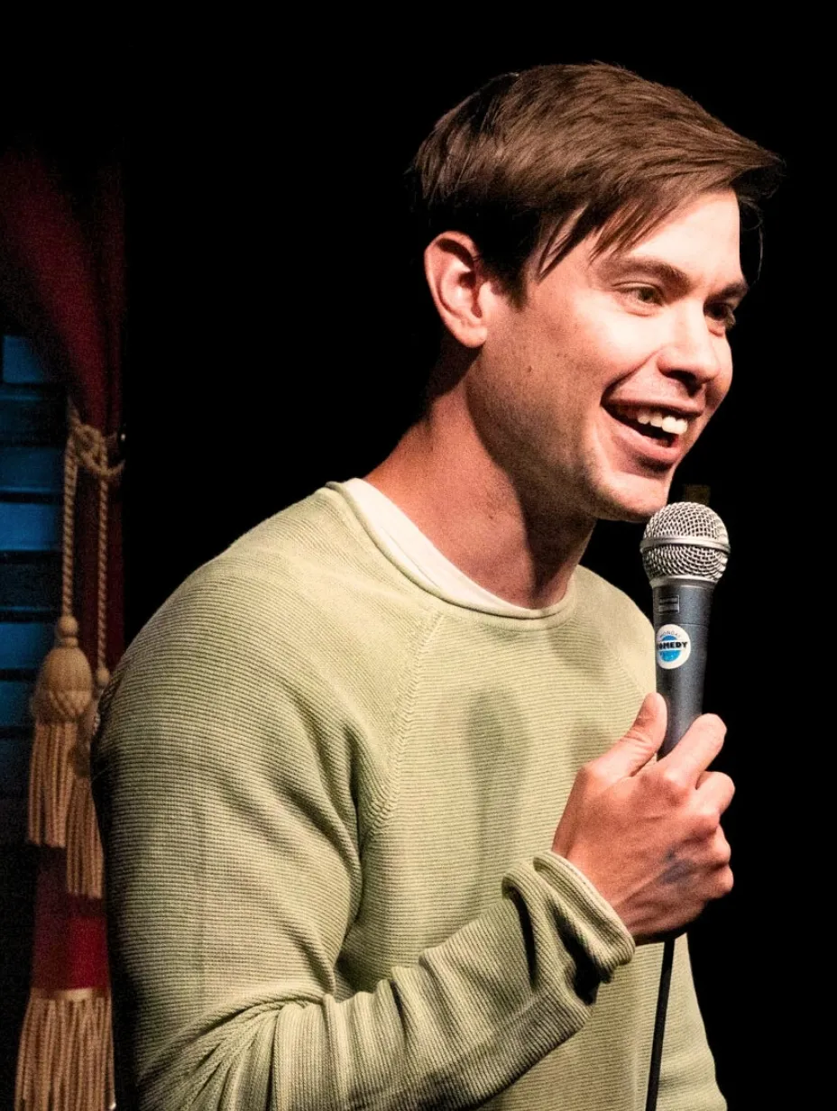Matt Foster, a male comedian, smiles widely and holds a microphone on stage, wearing a light-coloured crewneck jumper, ready to deliver his comedic treasure to a Comedy Freaks comedy show London.
