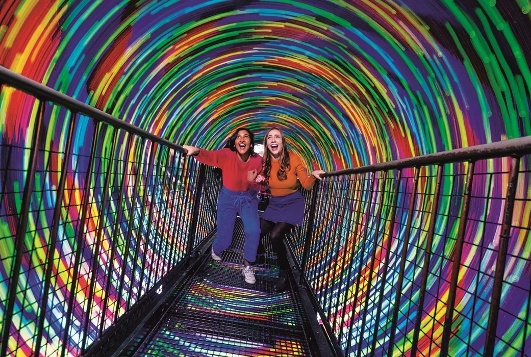 Two women laughing in colorful rainbow tunnel at Camera Obscura Edinburgh attraction.