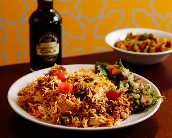A plate of biryani with a side salad sits on a wooden table, next to a bottle of Curiosity Cola and a bowl of curry. The background is an orange wall with a geometric pattern.