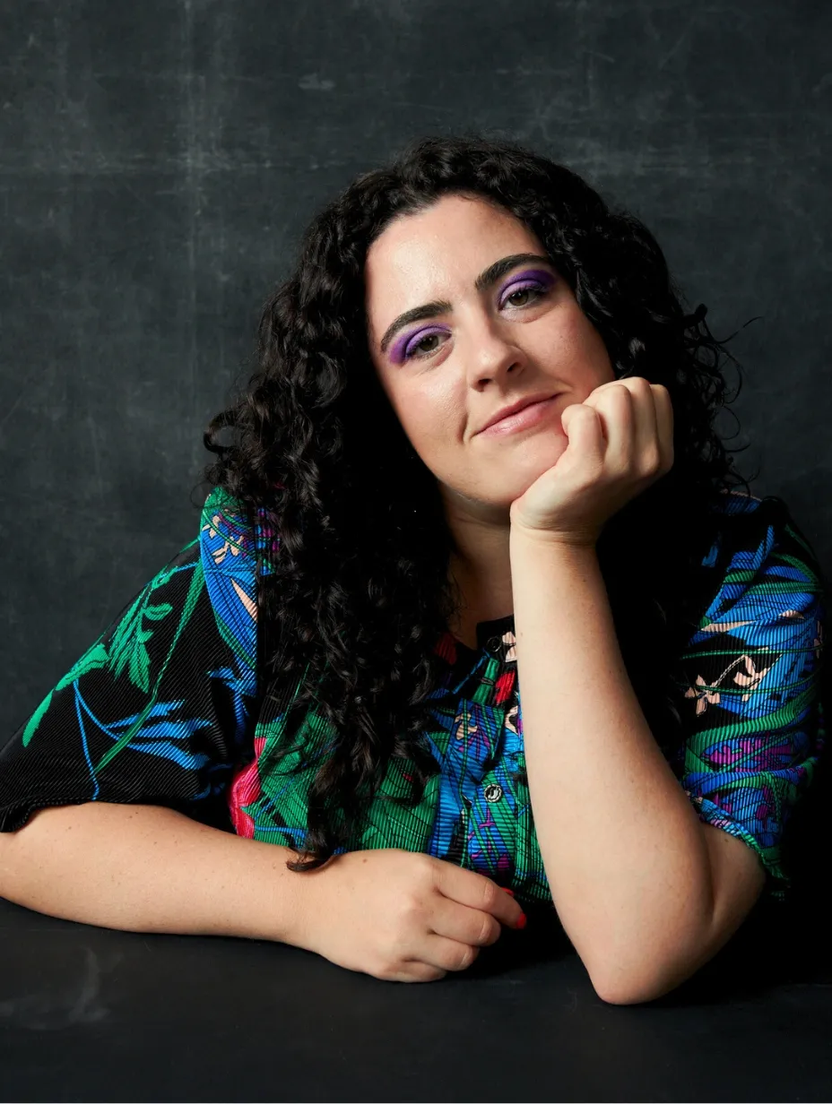 Jessica Fuchs, a female comedian, smiles confidently with her chin resting in her hand. She has long dark curly hair and is wearing a colourful shirt, embodying her razor-sharp and wildly relatable style at a Comedy Freaks comedy show London.