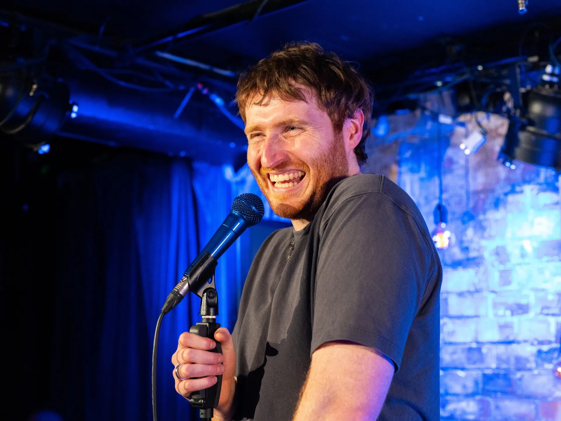 Ali Woods, a male comedian with short ginger hair and a beard, laughs heartily into a microphone on stage, with blue lighting and a brick wall backdrop at Comedy Freaks - the Wildest Comedy Club London.