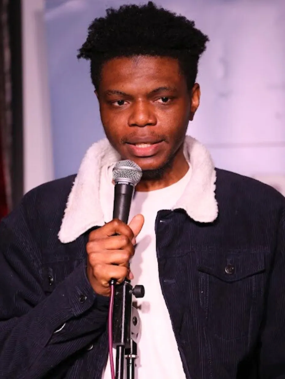 Onojie Idemudia, a male comedian, holds a microphone and looks at the audience from the stage, embodying his award-winning comedy and excellent delivery at a Comedy Freaks comedy show London.