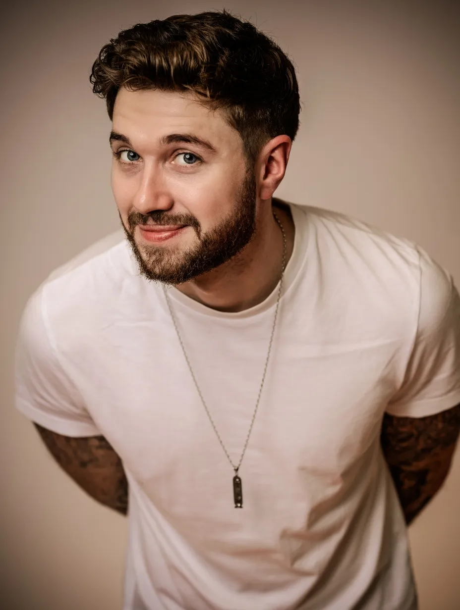 Joe Bingham, a male comedian with a beard and tattoos on his arms, smiles confidently at the camera while wearing a white t-shirt. He is a BBC New Comedy Award semi-finalist who is ready to deliver an unforgettable performance at a Comedy Freaks comedy show London.