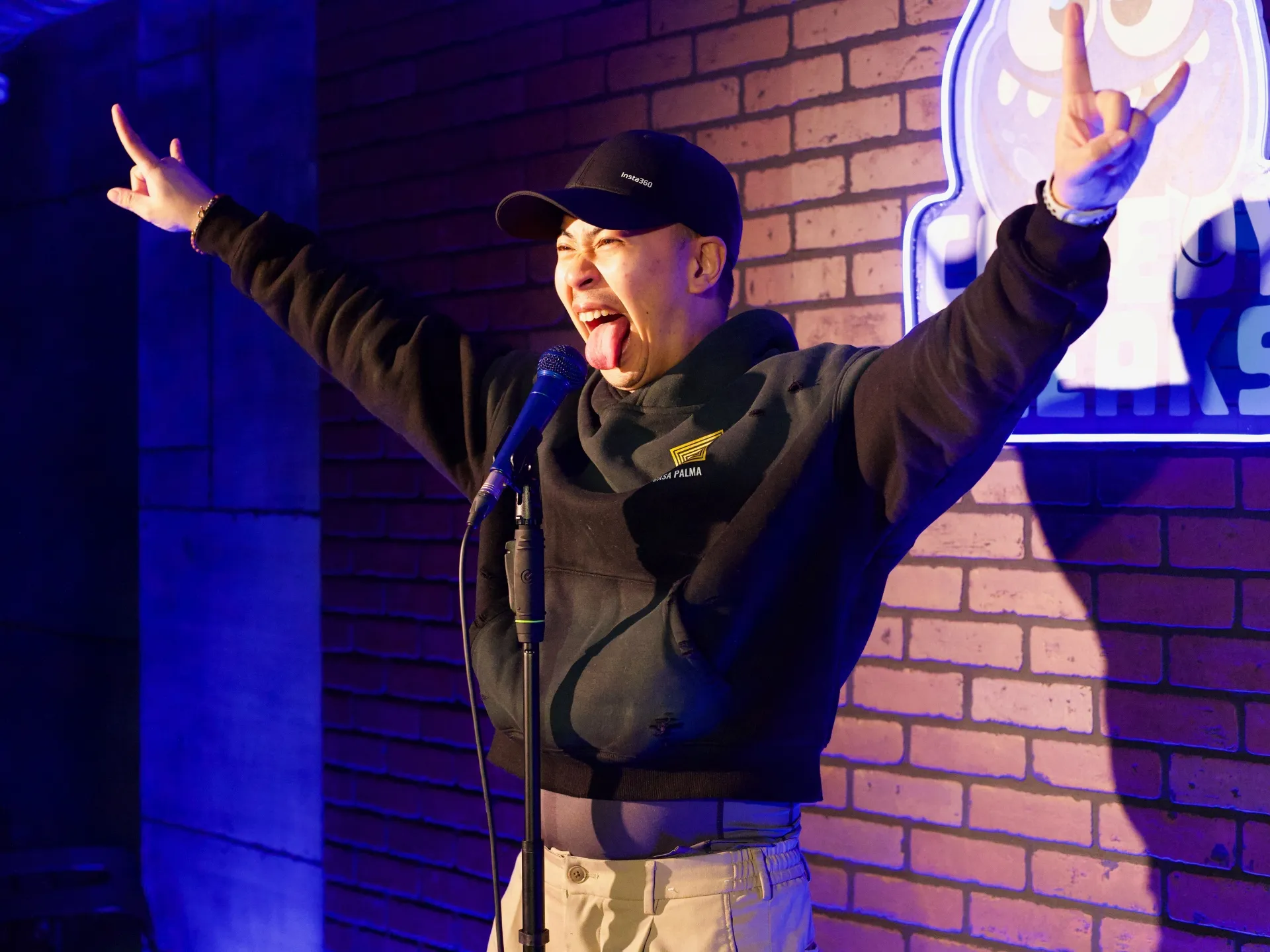 Lee J Ramirez, wearing a black cap and hoodie, enthusiastically throws his arms in the air and sticks out his tongue, on stage with the glowing Comedy Freaks logo and brick wall backdrop, in the best comedy club in London.