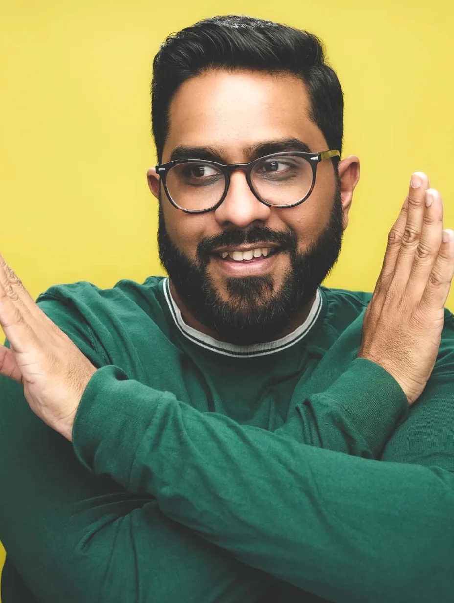 Eshaan Akbar, a male comedian with a beard and glasses, looks off to the side with a smirk while crossing his arms in an X shape, ready to deliver a sharp and insightful set at a Comedy Freaks comedy show London.
