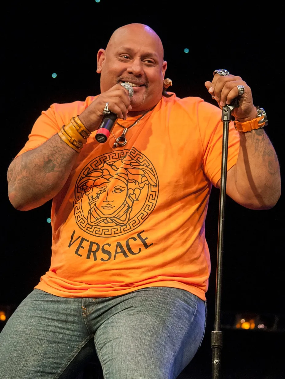 American comedian Dante Nero on stage in an orange Versace shirt, holding a microphone and a stand, a performer in the London stand-up comedy scene.