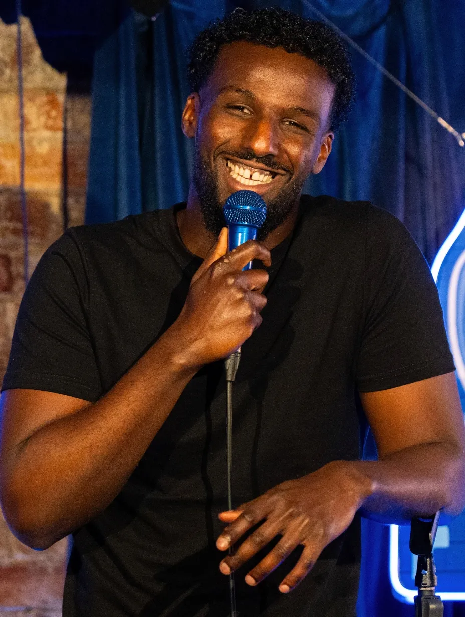 Bamph, a comedian, smiles broadly as he holds a microphone, poised for his performance.