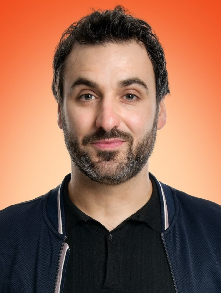 A close-up headshot of comedian Patrick Monahan with a beard, a black polo shirt, and a dark blue jacket, a performer in the London comedy scene.