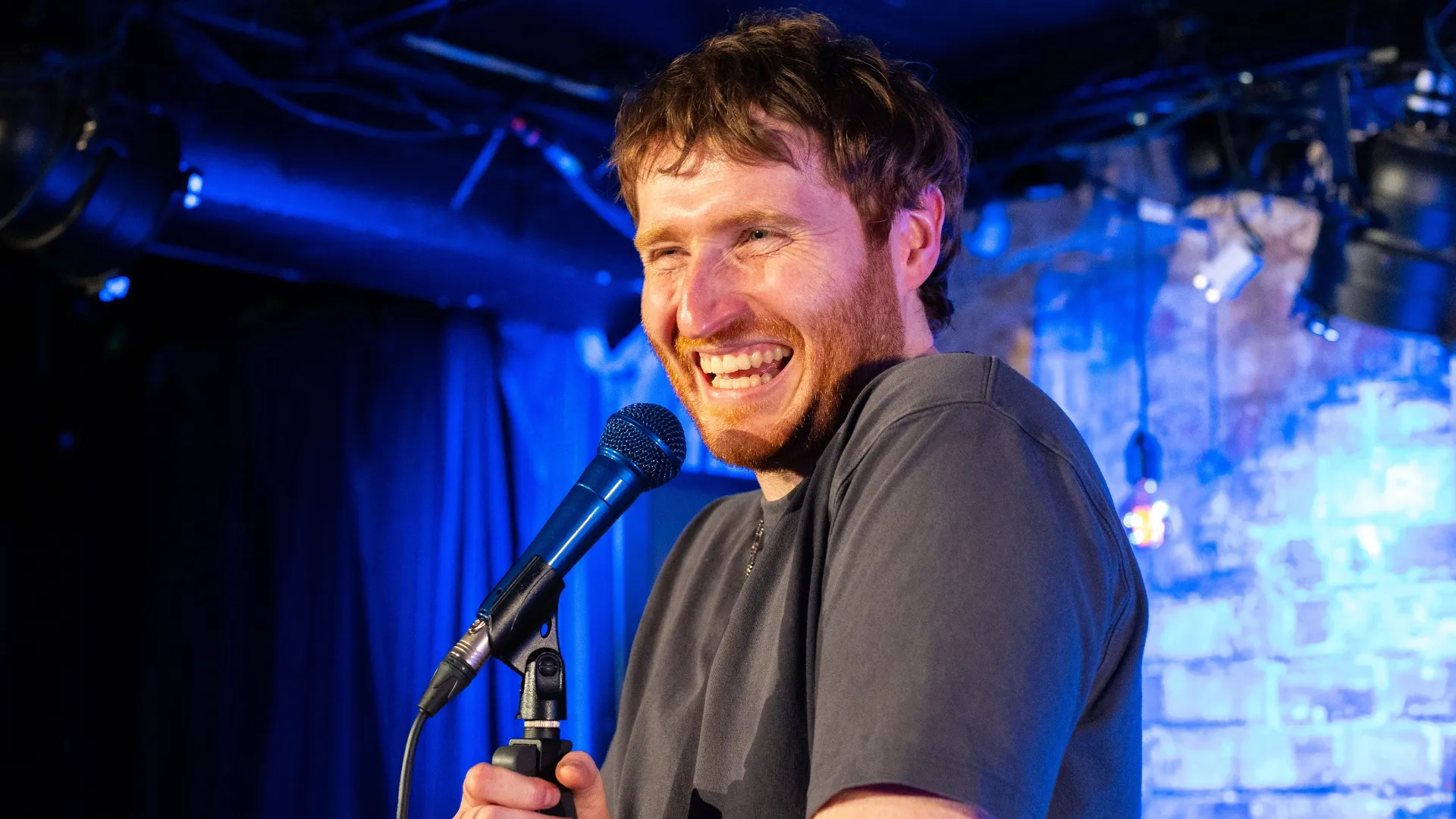 Ali Woods, a male comedian with short ginger hair and a beard, laughs heartily into a microphone on stage, with blue lighting and a brick wall backdrop at Comedy Freaks - the Wildest Comedy Club London.