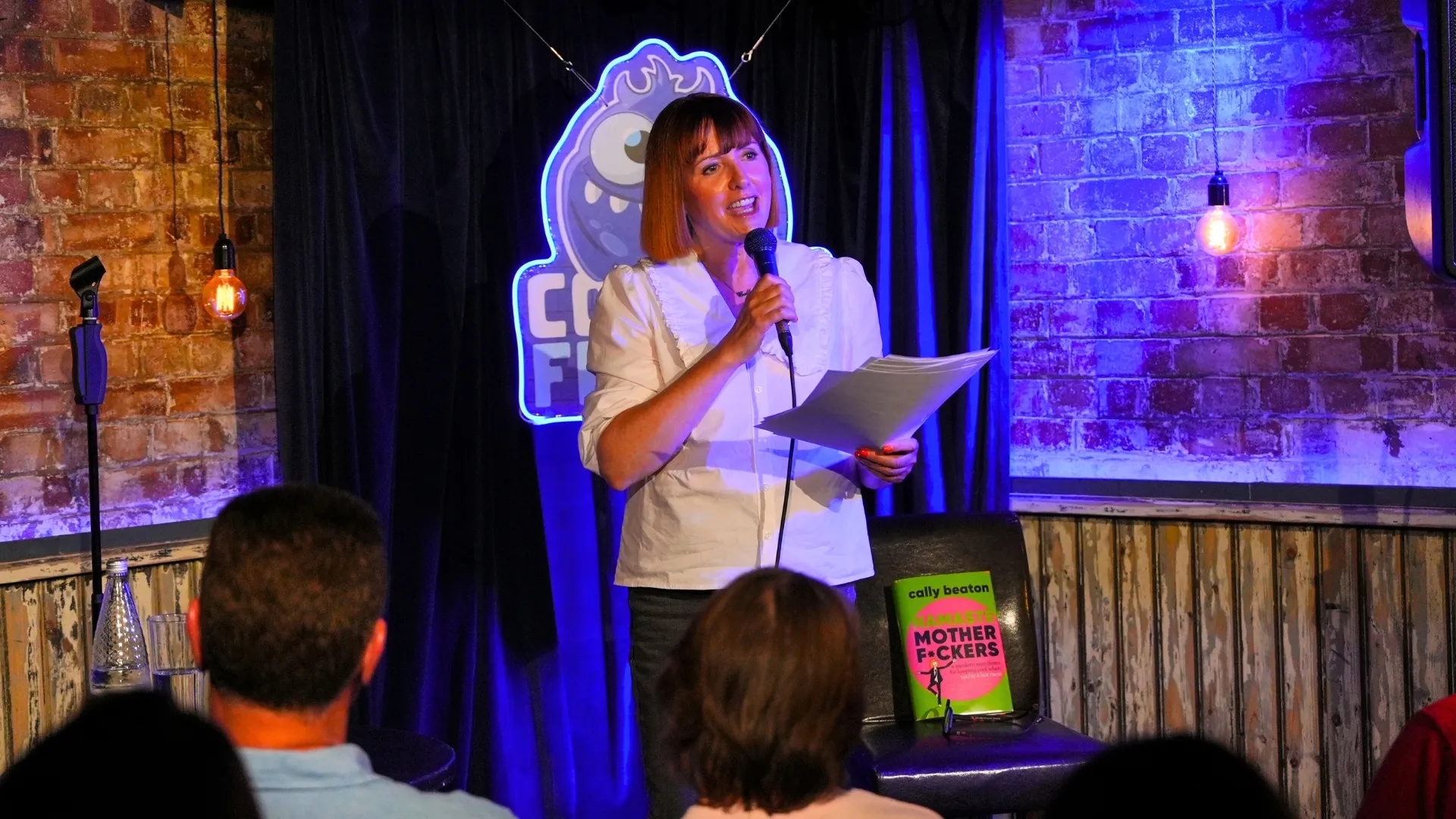 Cally Beaton, a female comedian with ginger hair, stands on stage holding a microphone, engaging with the audience in front of blue curtains and brick walls, delivering a captivating performance at the best comedy show in London -Comedy Freaks.