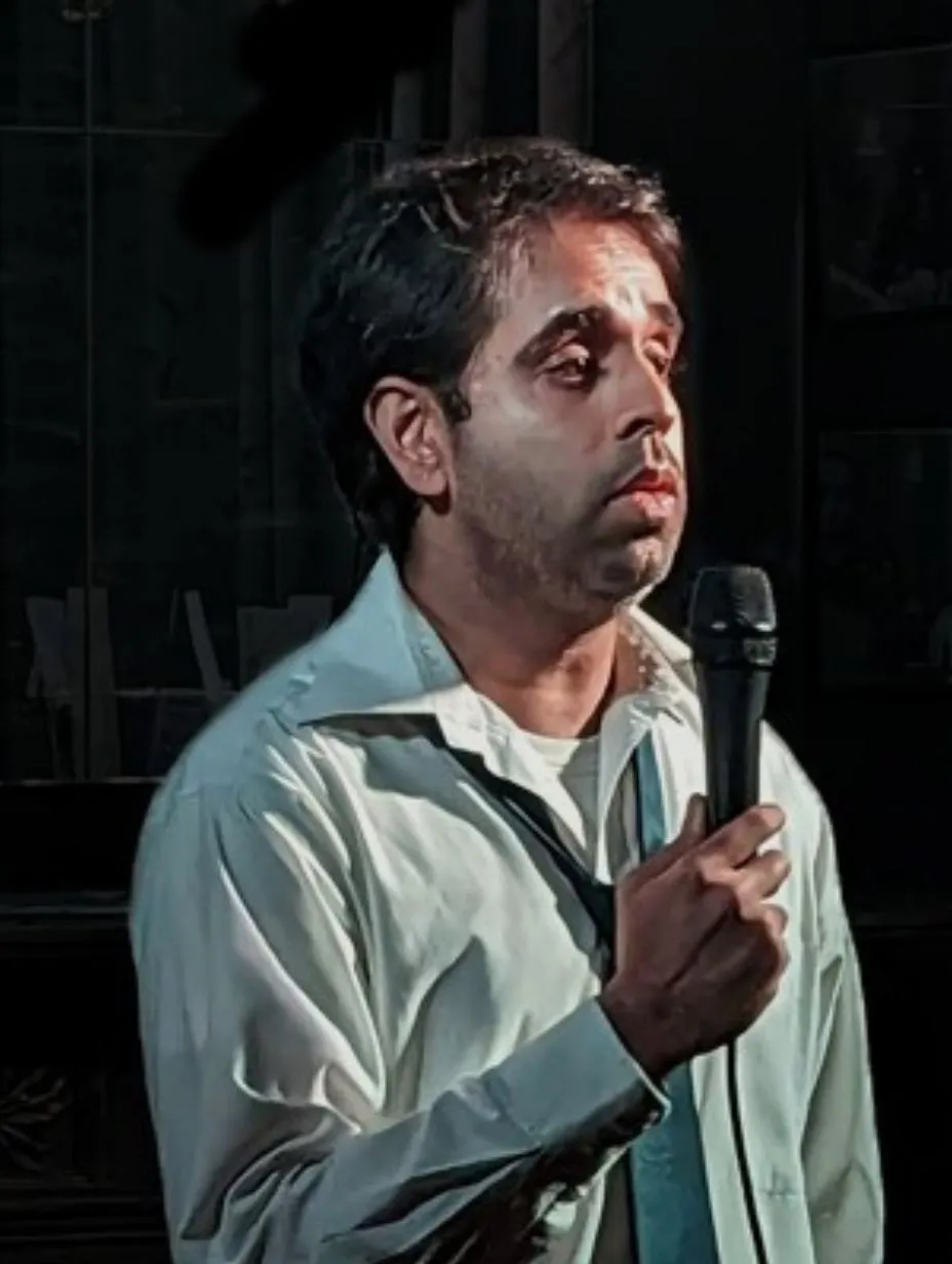 Portrait of comedian Mahesh Khilnani holding a microphone on stage. He is wearing a light-coloured, slightly rumpled dress shirt and a loosened tie against a dark background. Catch this rising UK comedian at a comedy club London.