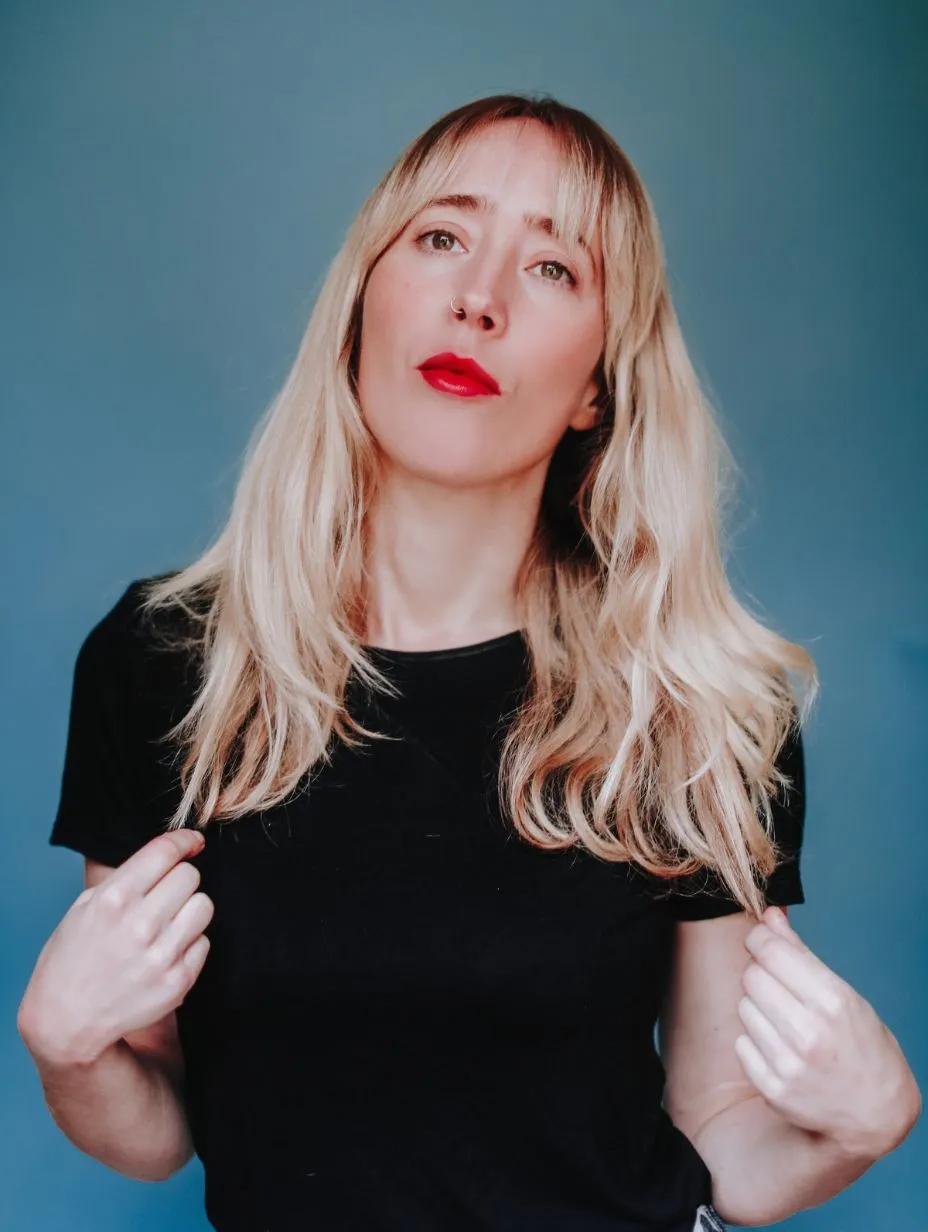 Portrait of comedian Kate-Lois Elliott with long blonde hair and striking red lipstick, wearing a black t-shirt against a blue background. This is one of the best comedians performing in London.