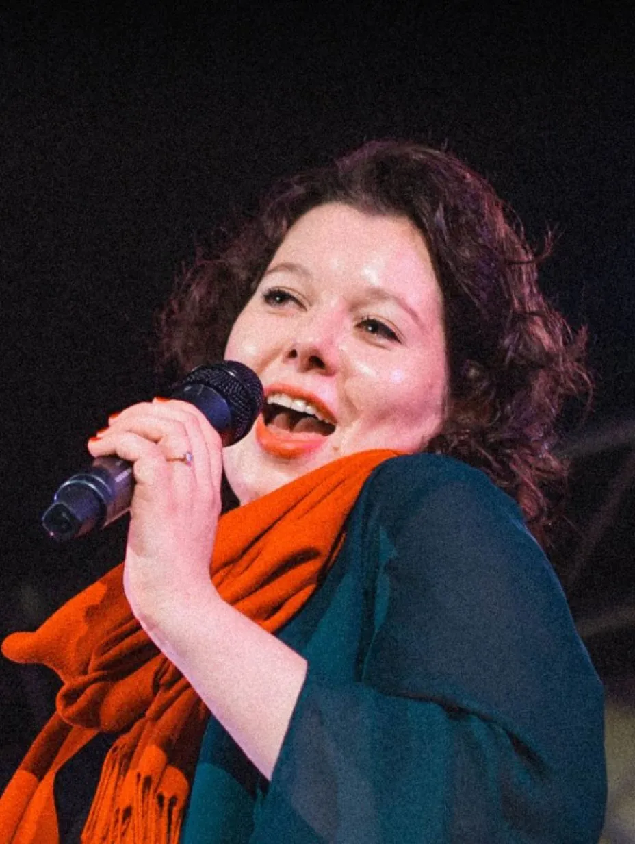 Close up of comedian Mara Mainka on stage, singing or laughing passionately into a microphone while wearing a dark top and a vibrant orange scarf against a dark background. This performer is part of London comedy shows.