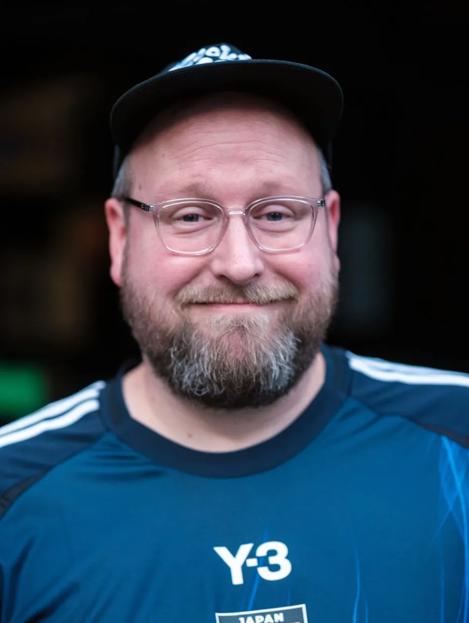 Close-up portrait of comedian Lee Hudson smiling. He has a full beard, wears clear-rimmed glasses, a black baseball cap backwards, and a blue Adidas Y-3 football jersey featuring the Japan crest. This is one of the best comedians performing comedy in London.
