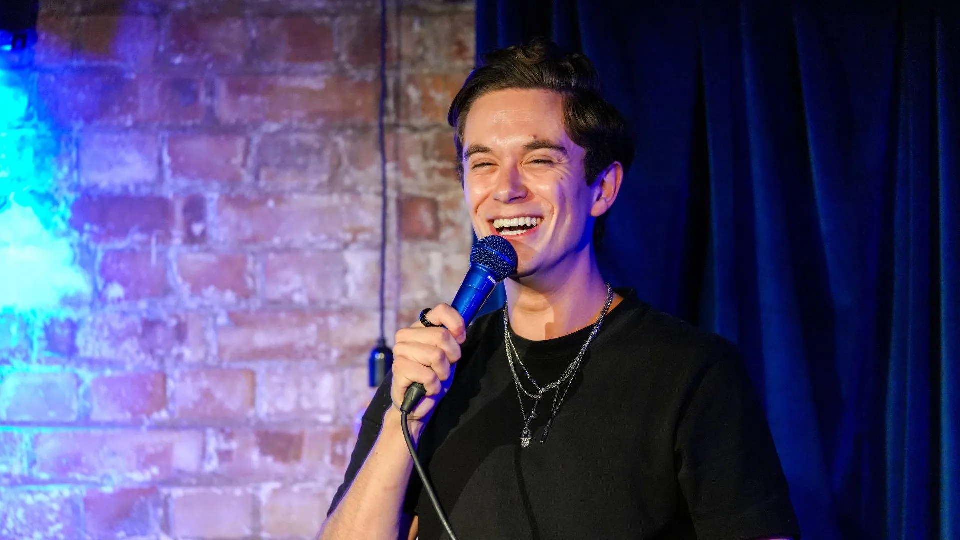 Jomi Cruz, a male comedian with dark hair, laughs while holding a microphone on stage against a brick wall with blue lighting, delivering an energetic and wild performance at Comedy Freaks, most unique comedy show London.