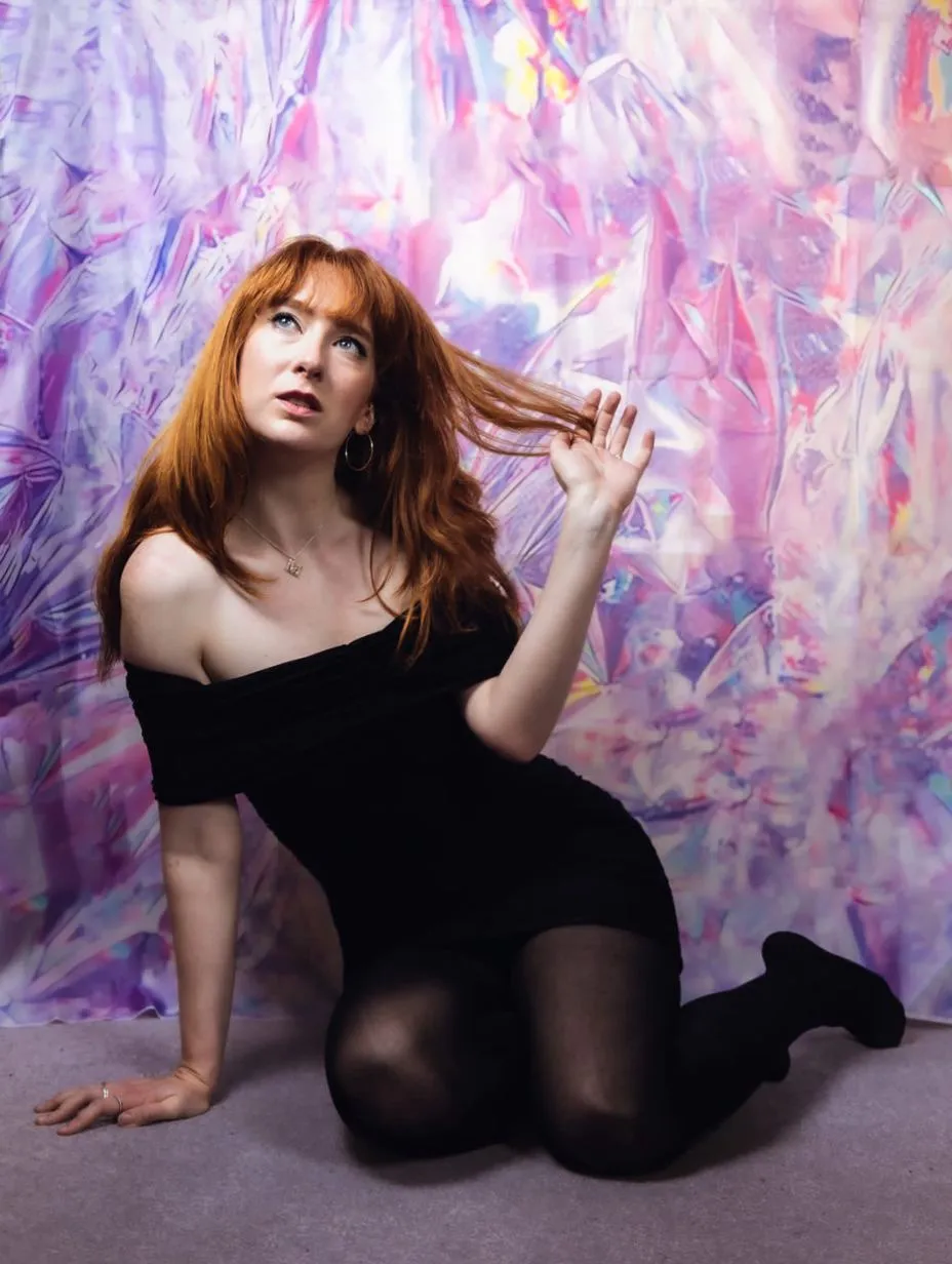 Portrait of comedian Madeleine Munford with long red hair and a black off the shoulder dress, posing against a vibrant, iridescent background. This comedian brings some of the best stand up comedy London has to offer.